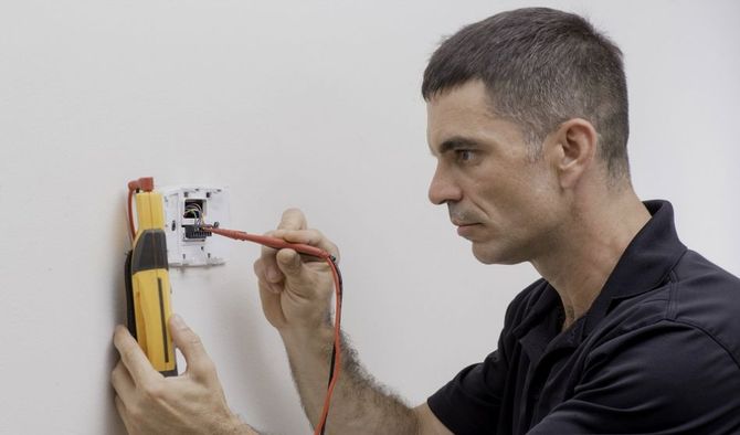 A professional uses a multimeter to test electrical wiring on a white wall.