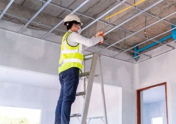 A worker in a high-visibility vest and hard hat stands on a ladder, installing electrical wires in a metal ceiling frame.