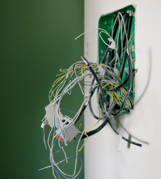 Exposed electrical wiring hanging from a wall box on a white wall next to a green wall.