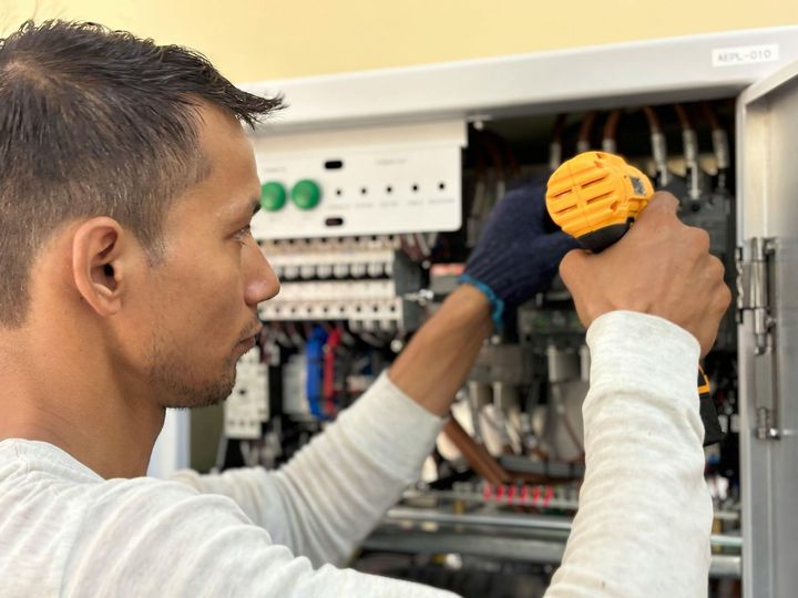 An electrician wearing a plaid shirt and blue overalls kneels to install or repair a wall outlet.