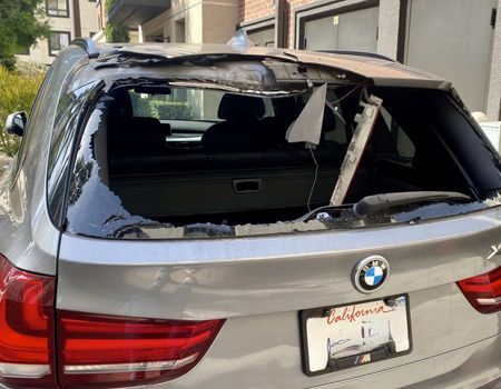 Silver BMW SUV with shattered rear windshield and hanging broken glass in a driveway