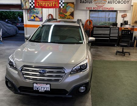Silver Subaru SUV in an auto body shop, facing forward with a Ferrari sign and tool cabinets in the background