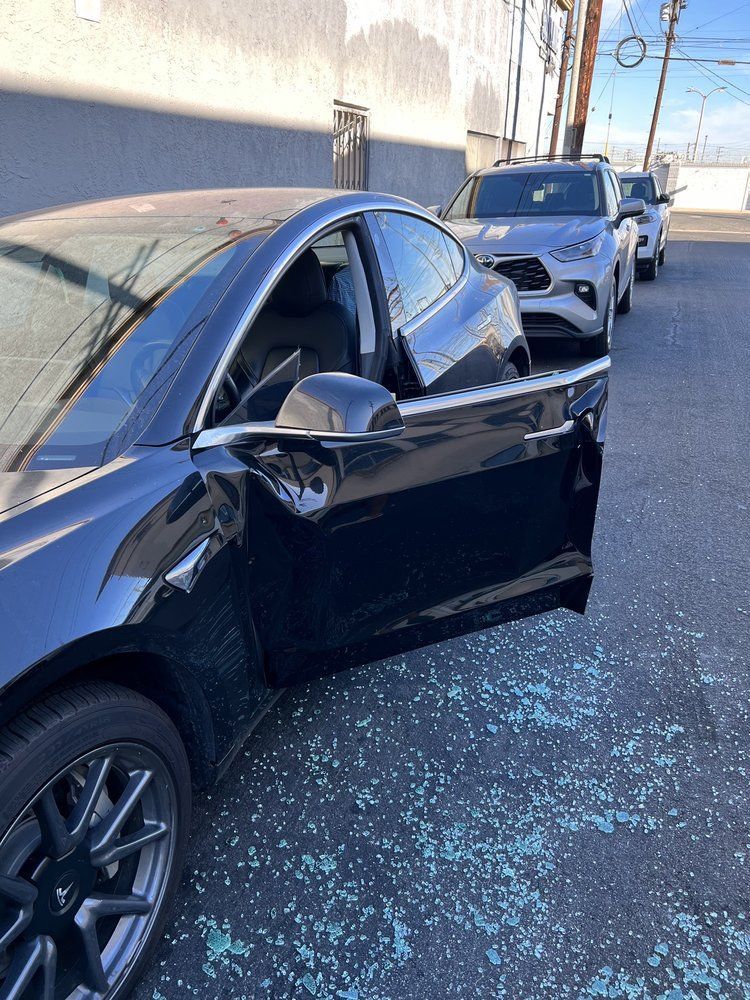 Black sedan with shattered windows and broken glass on the ground, parked along a wall with another car behind it