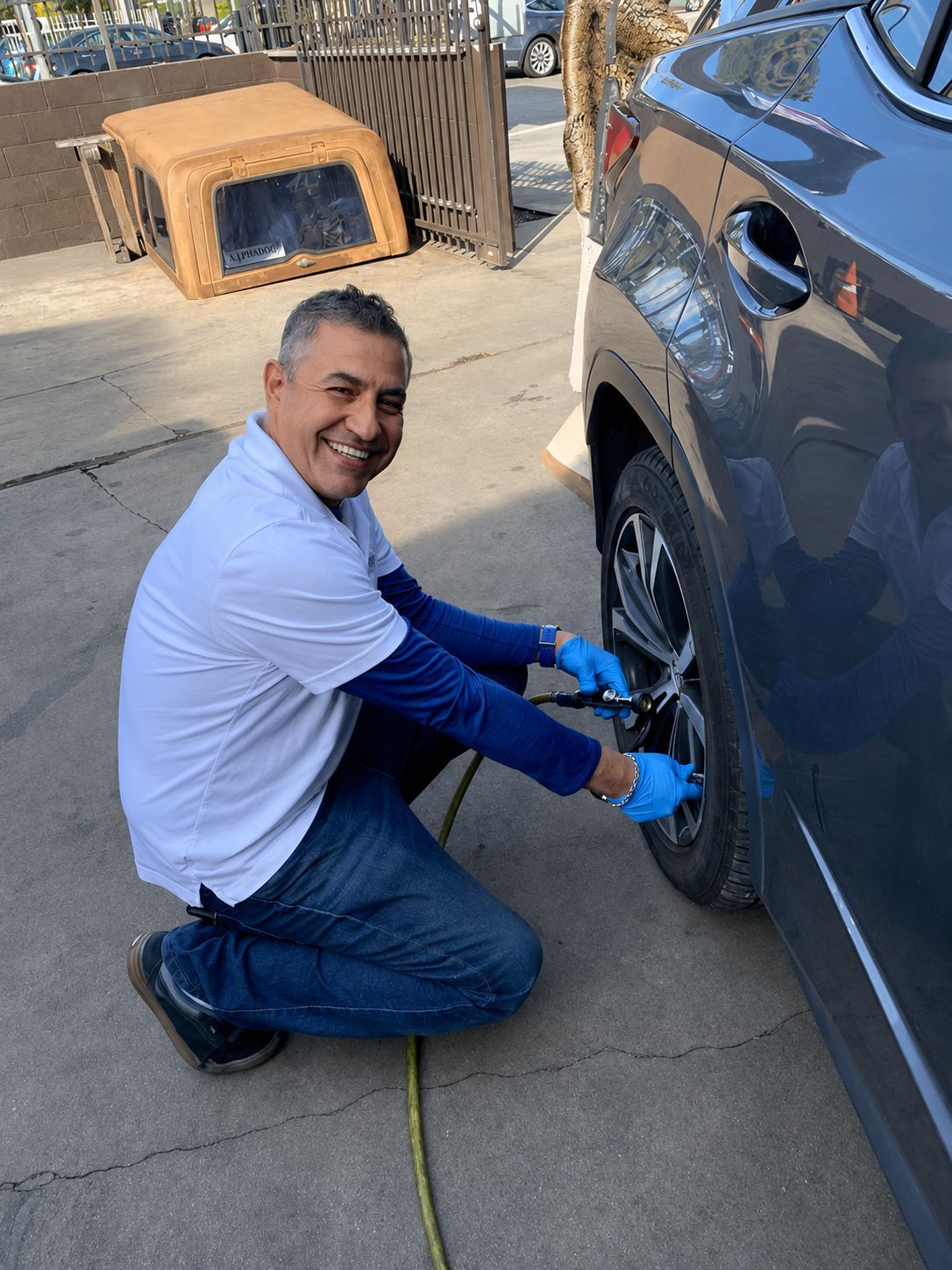 Man kneels beside a dark SUV, smiling while using an air hose to fill a tire.