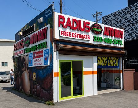 Corner auto body shop with red and green signage, glass storefront, and open garage bay on a sunny street