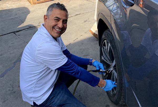 Man in blue gloves crouches beside a car, working on the rear wheel and smiling at the camera.