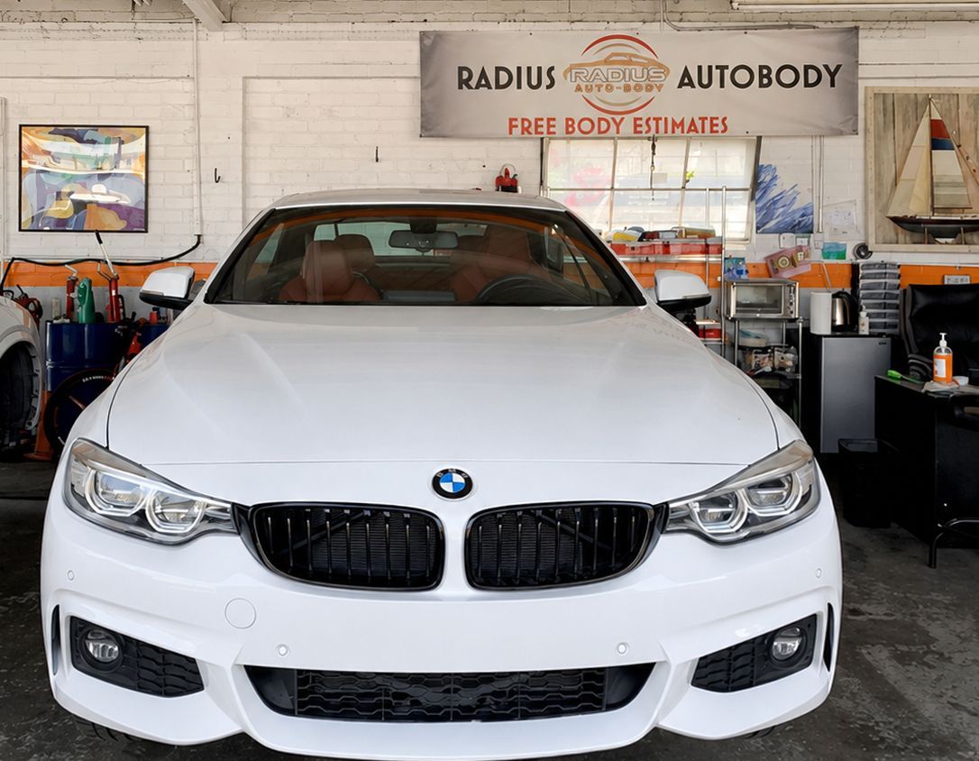 White BMW sedan in an auto body shop garage with tools and a “Radius Autobody” sign.