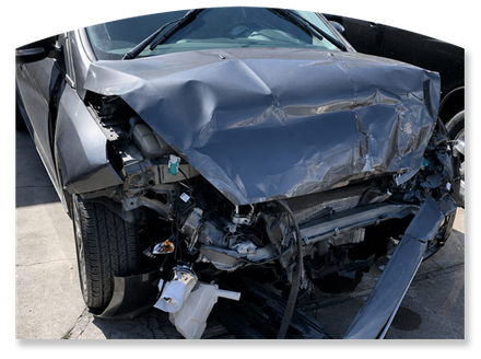 Severely damaged front end of a dark gray car after a collision, with crumpled hood and exposed wheel