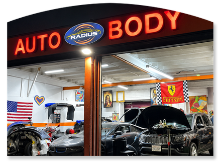 Auto body shop at night with cars inside, lit by neon “AUTO BODY” sign.