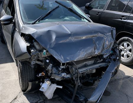 Front of a black car heavily damaged in a collision, with the hood crumpled and bumper torn off.