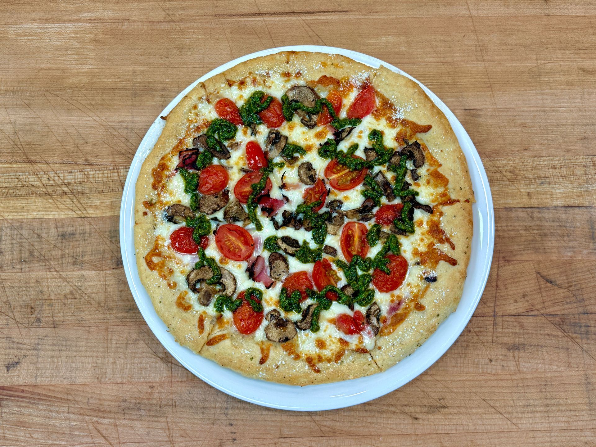 Pizza on white plate, topped with tomatoes, mushrooms, and pesto, on a wooden table.