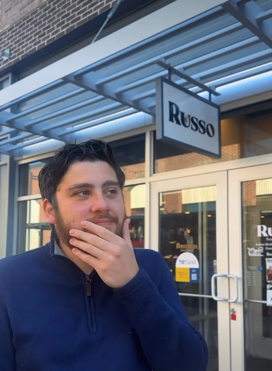 Man in blue sweater, hand to chin, outside Russo storefront.