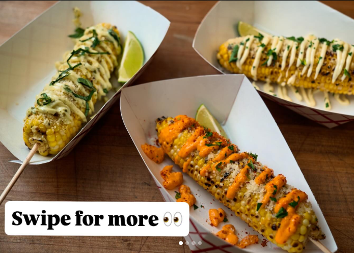 Three ears of corn on the cob, dressed with sauces, herbs, and lime wedges in paper trays on wood.