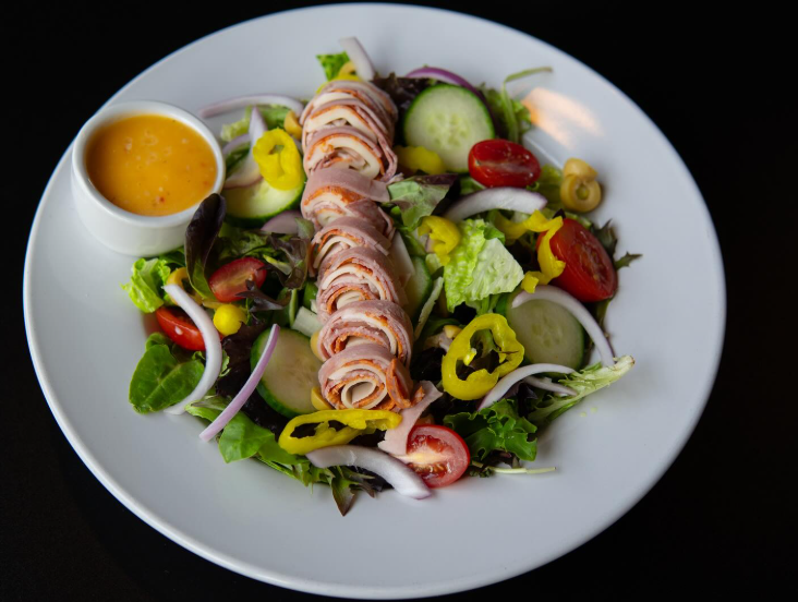 Salad with ham roll, mixed greens, tomatoes, cucumbers, onions, peppers, and dressing in a white bowl.