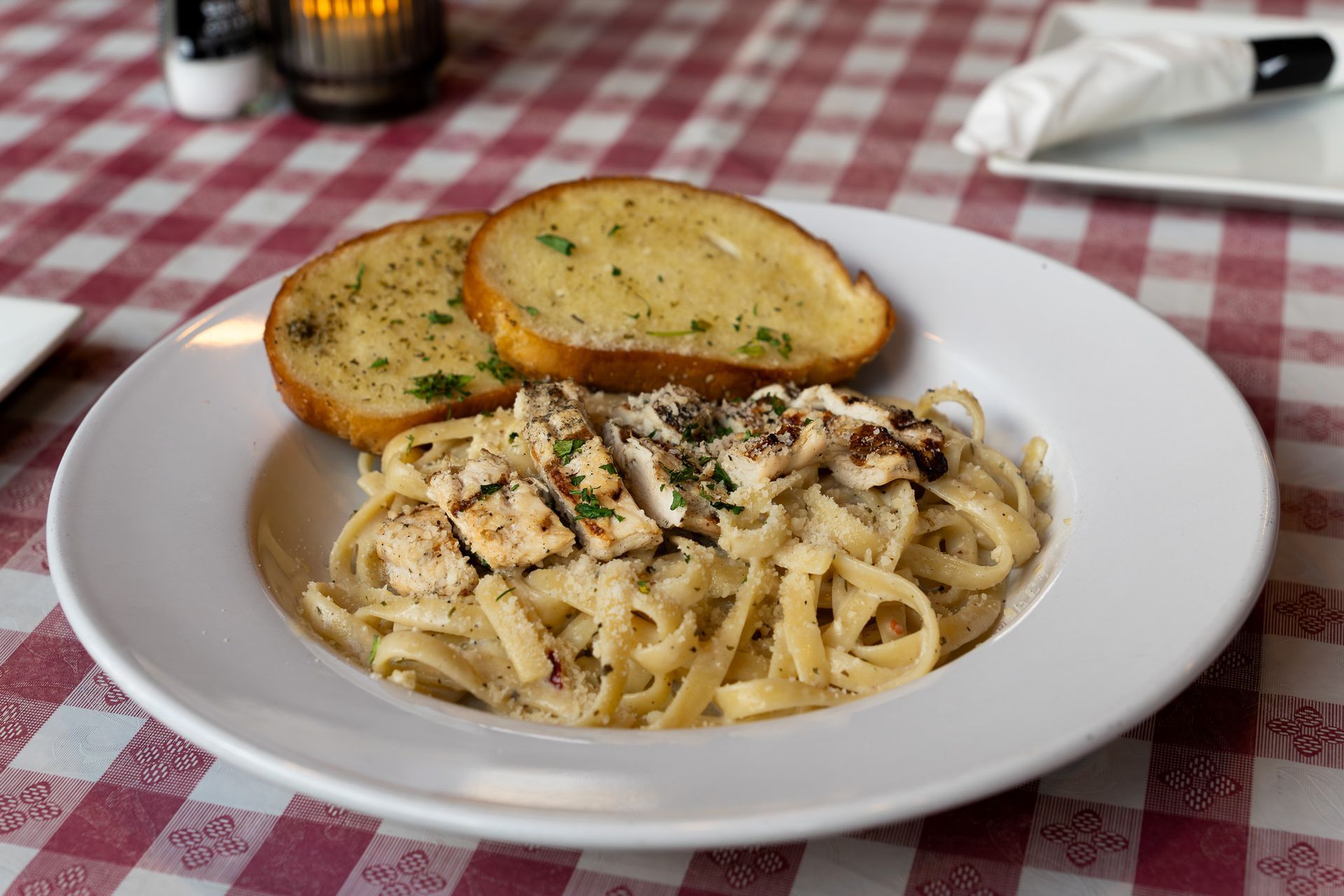 Fettuccine Alfredo with grilled chicken and garlic bread on a white plate, red checkered tablecloth.