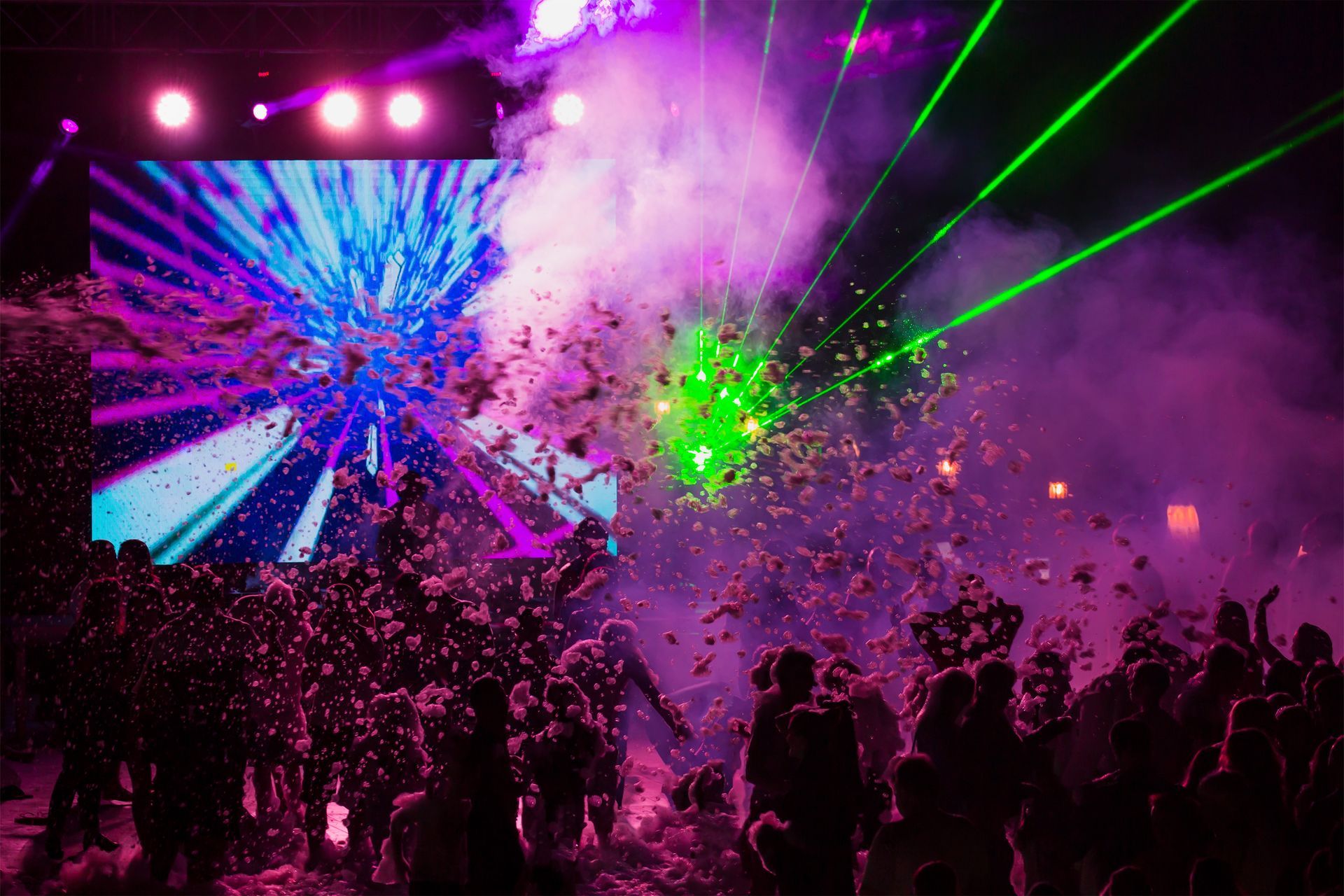 A vibrant club scene with lasers, fog, and a crowd dancing under a colorful screen.