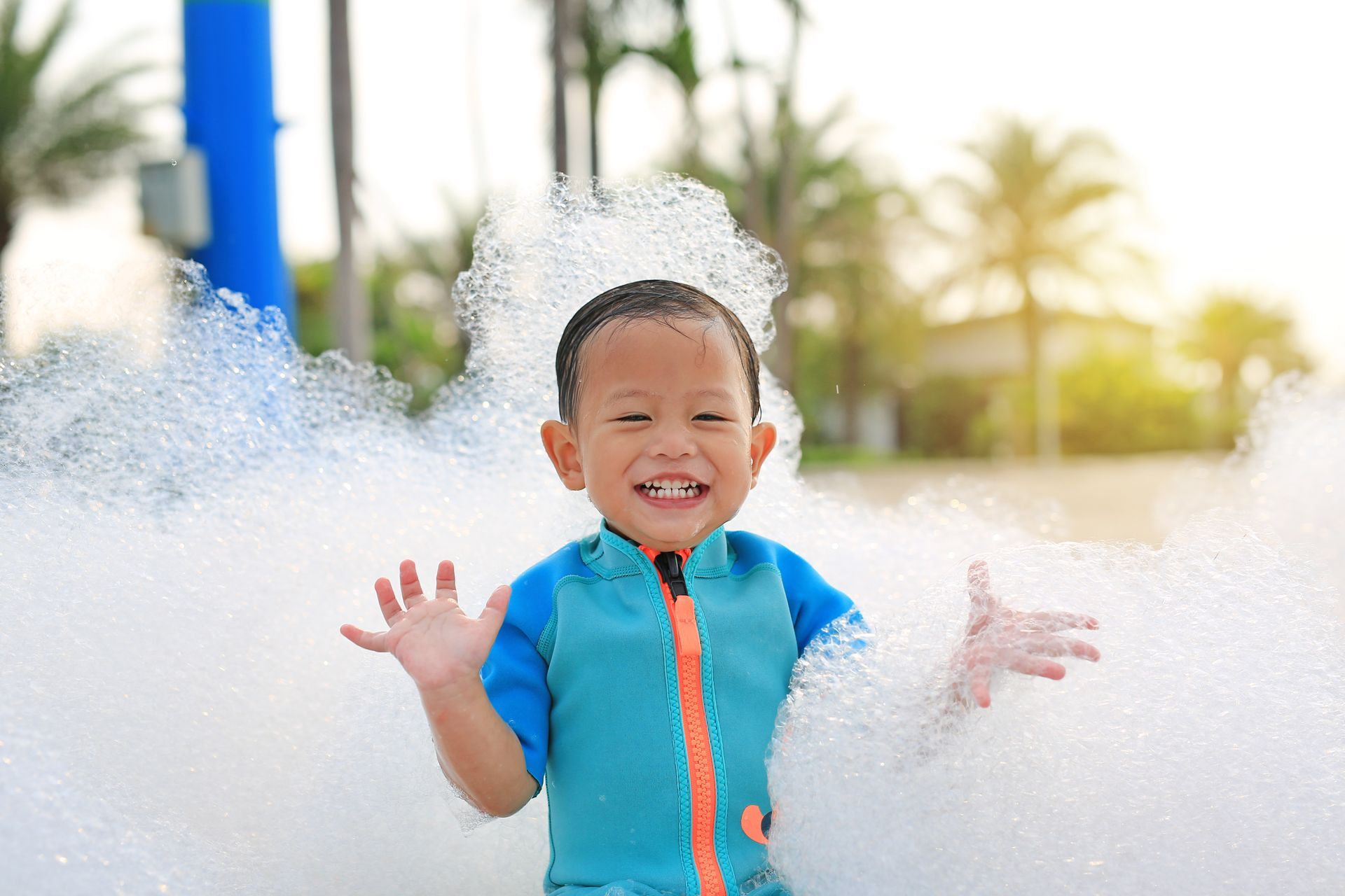 Young boy in blue swimsuit smiles in a sea of white bubbles; outdoors, sunny.