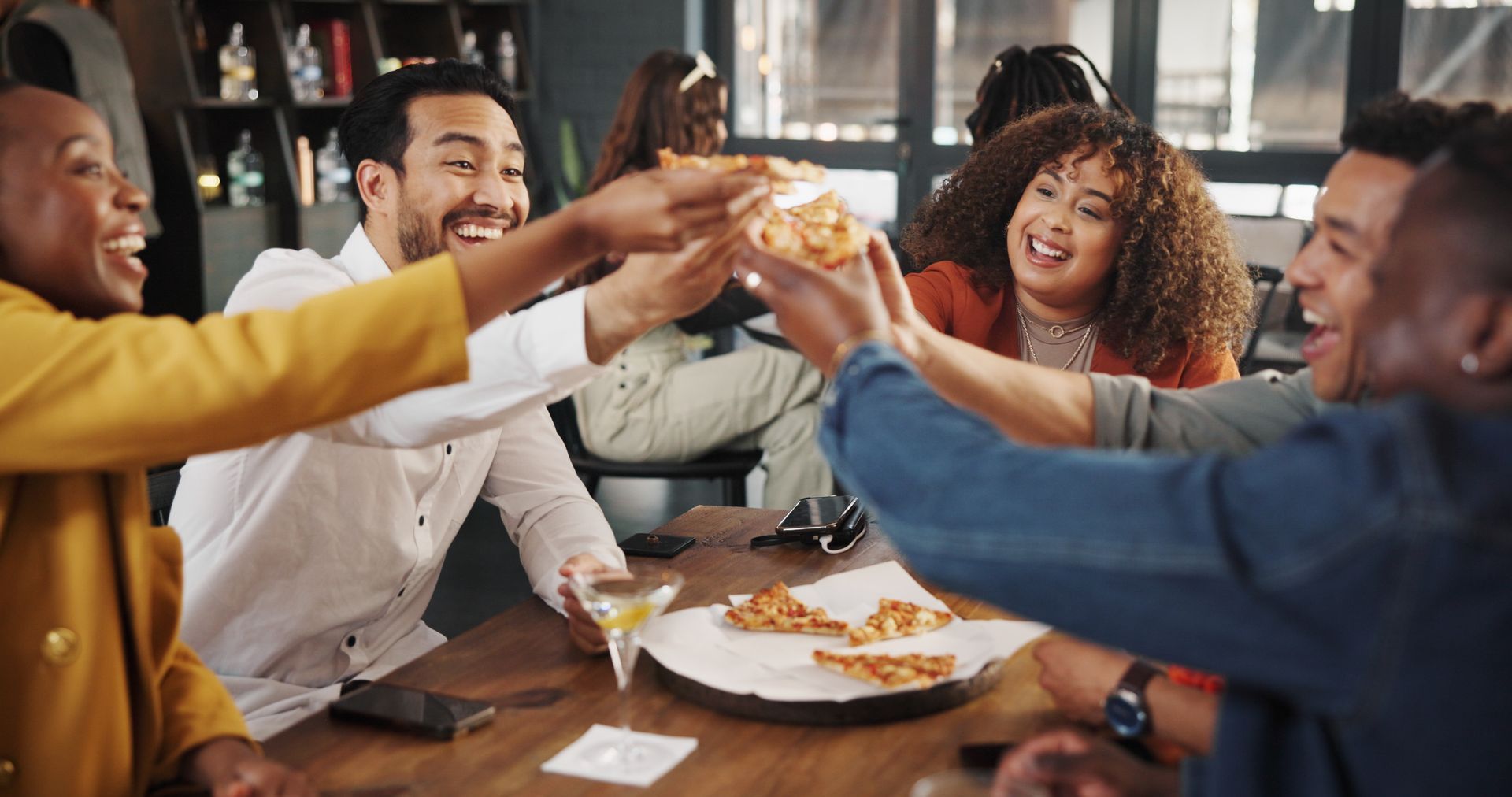 A close-up shot of a group of cheerful friends gathered around a wooden table at a restaurant, raising slices of pizza together in a celebratory toast. A man on the left and a woman with curly hair on the right look on, smiling and laughing, while additional pizza slices rest on a tray in the center of the table.