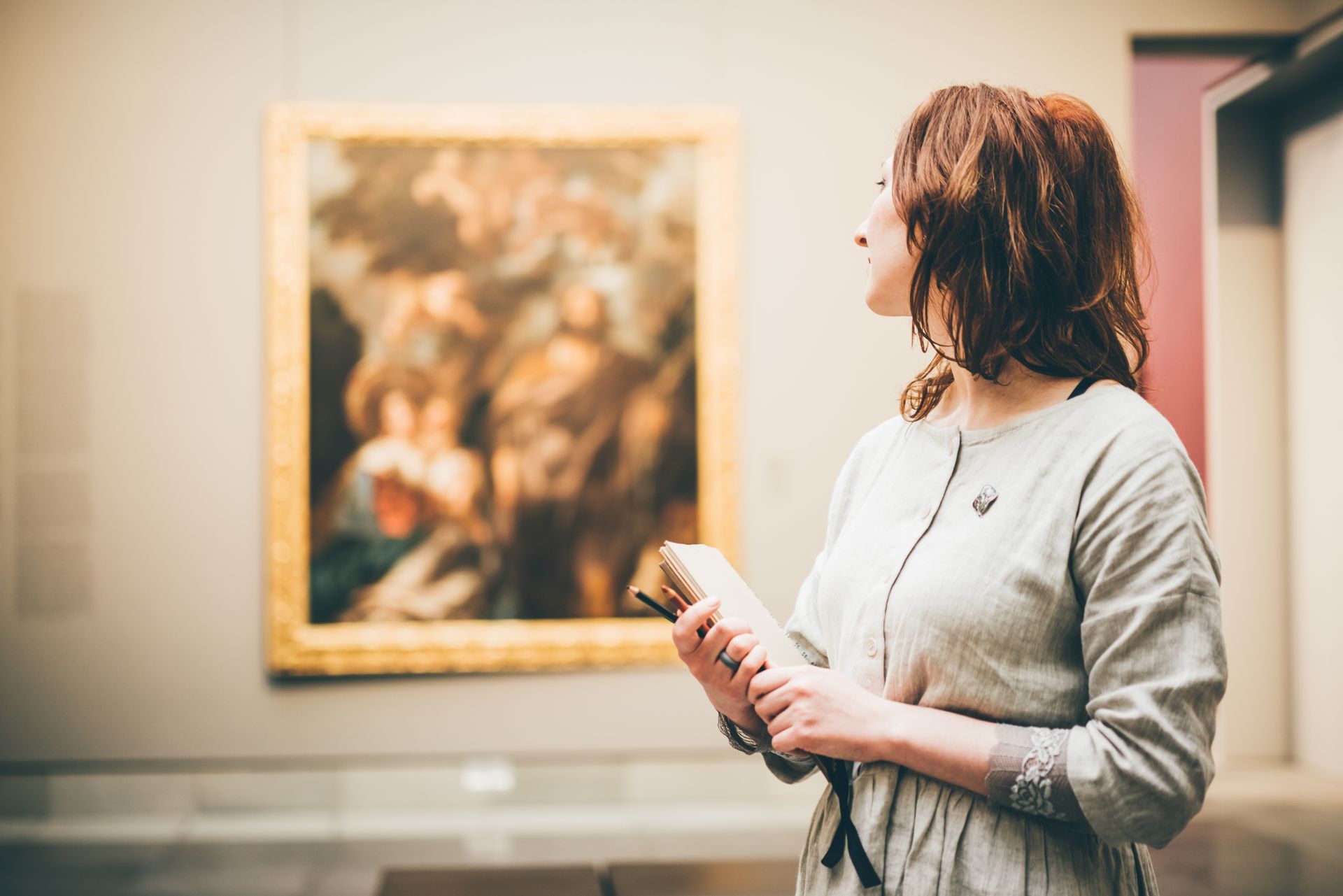 Alt Text: A side profile shot of a young woman with brown hair holding a notebook and a pen inside an art museum. She stands in the foreground, looking toward a large, gold-framed classical painting hanging on the wall in the blurred background.