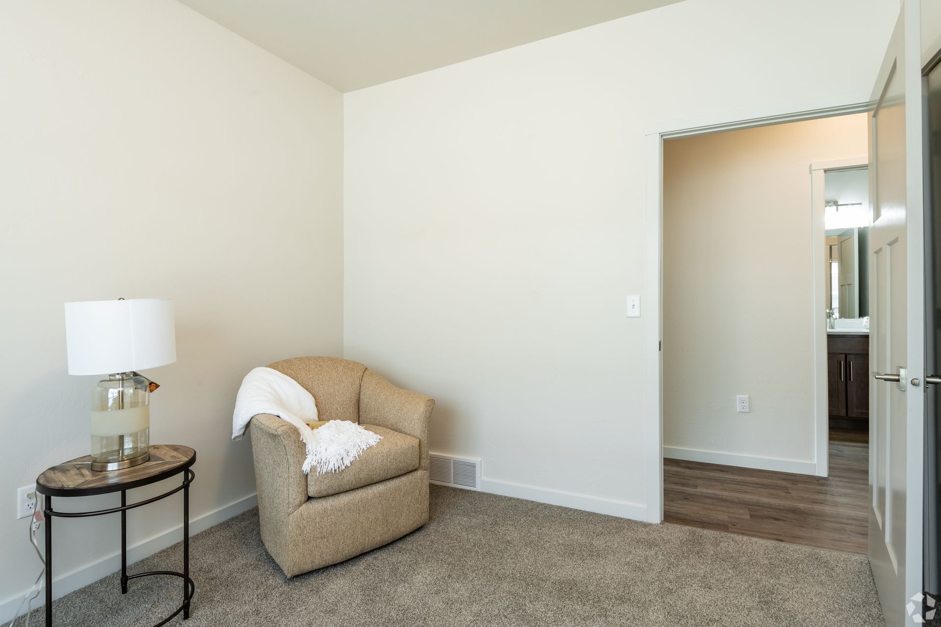 Empty beige room with a chair, side table, lamp, and doorway to a hallway and kitchen.