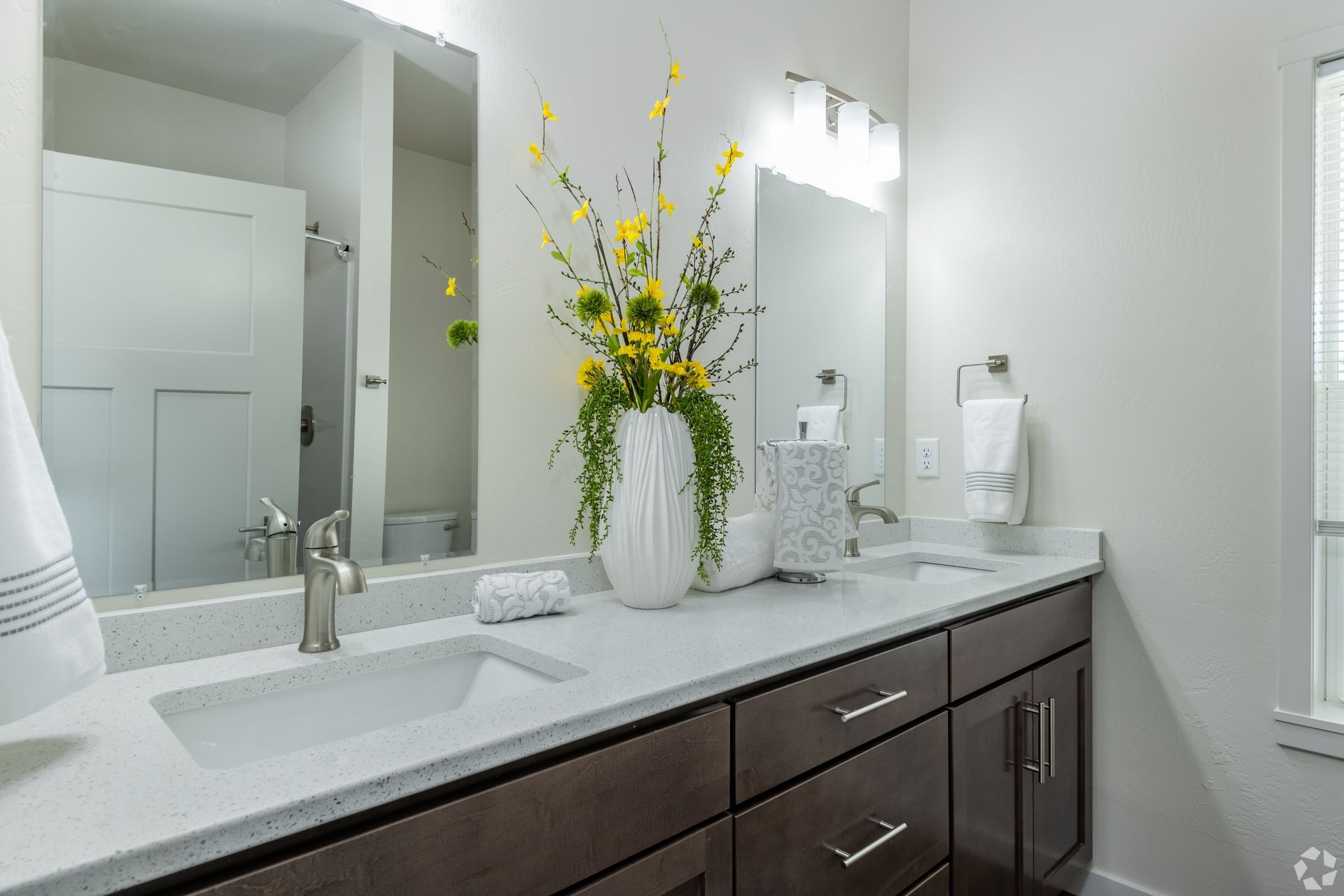 Modern bathroom vanity with white countertop, dark wood cabinets, mirror, and a white vase of yellow flowers