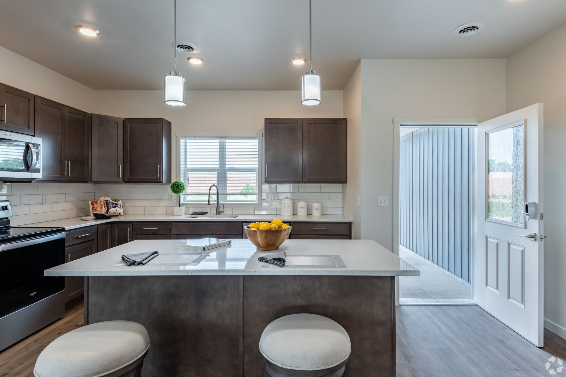 Bright modern kitchen with island, pendant lights, dark cabinets, and open door to a laundry room.