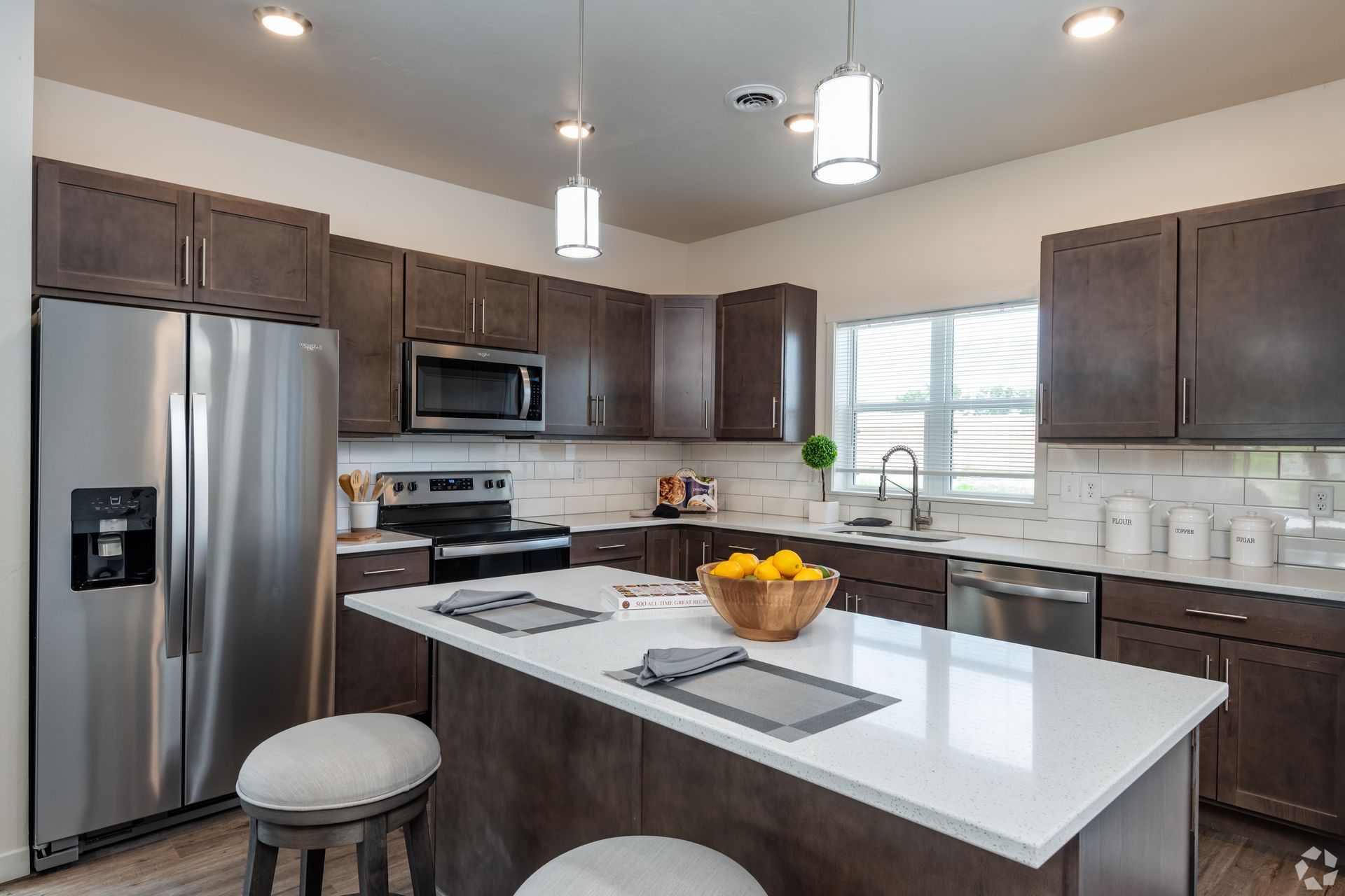 Modern kitchen with stainless steel appliances, dark cabinets, white island, and bowl of fruit