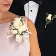 Formal couple with white floral boutonnieres and corsage, man in black tuxedo and woman in pale pink dress.