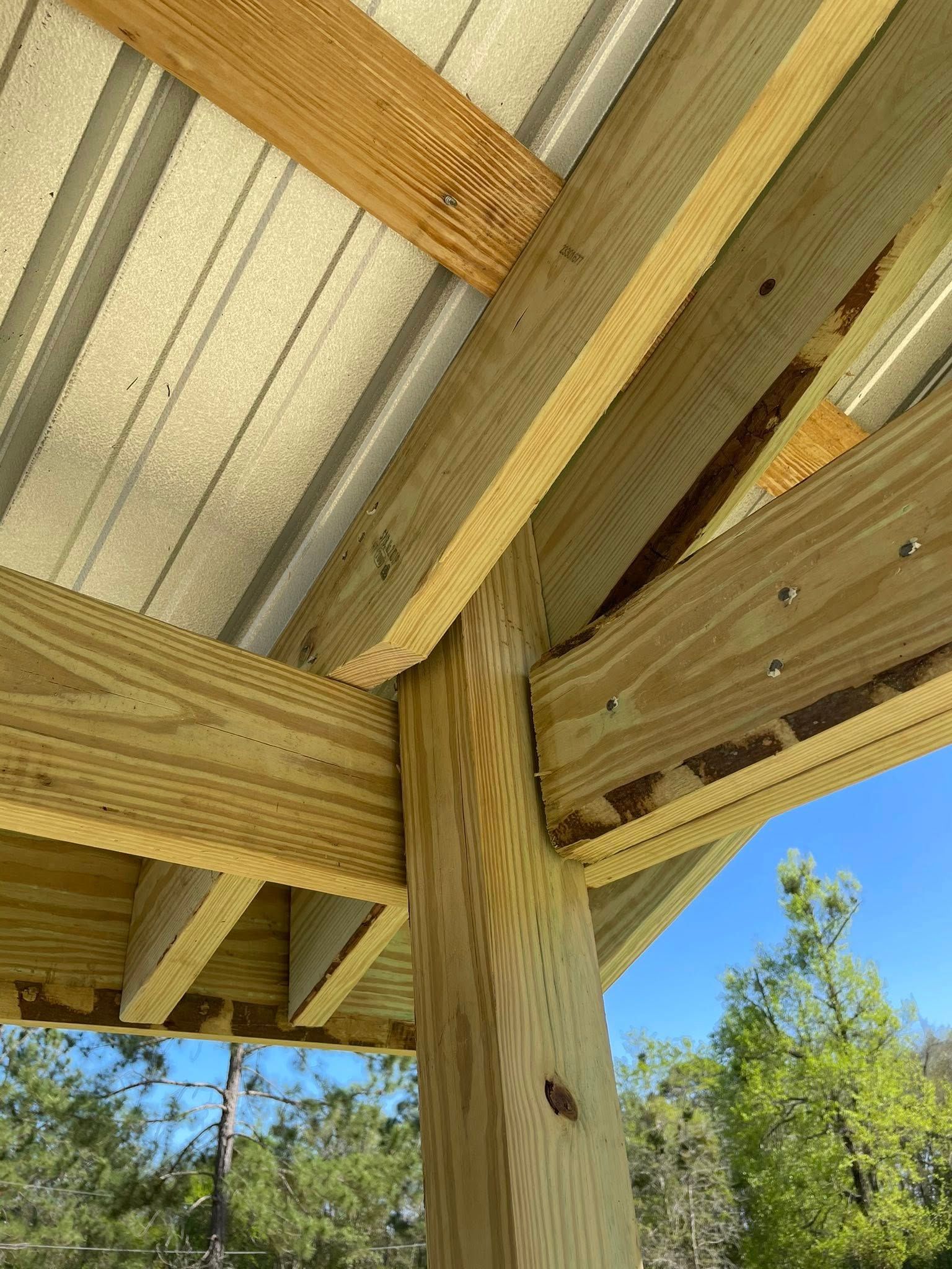 Close-up of a wooden deck support beam and roof underside against blue sky and trees.
