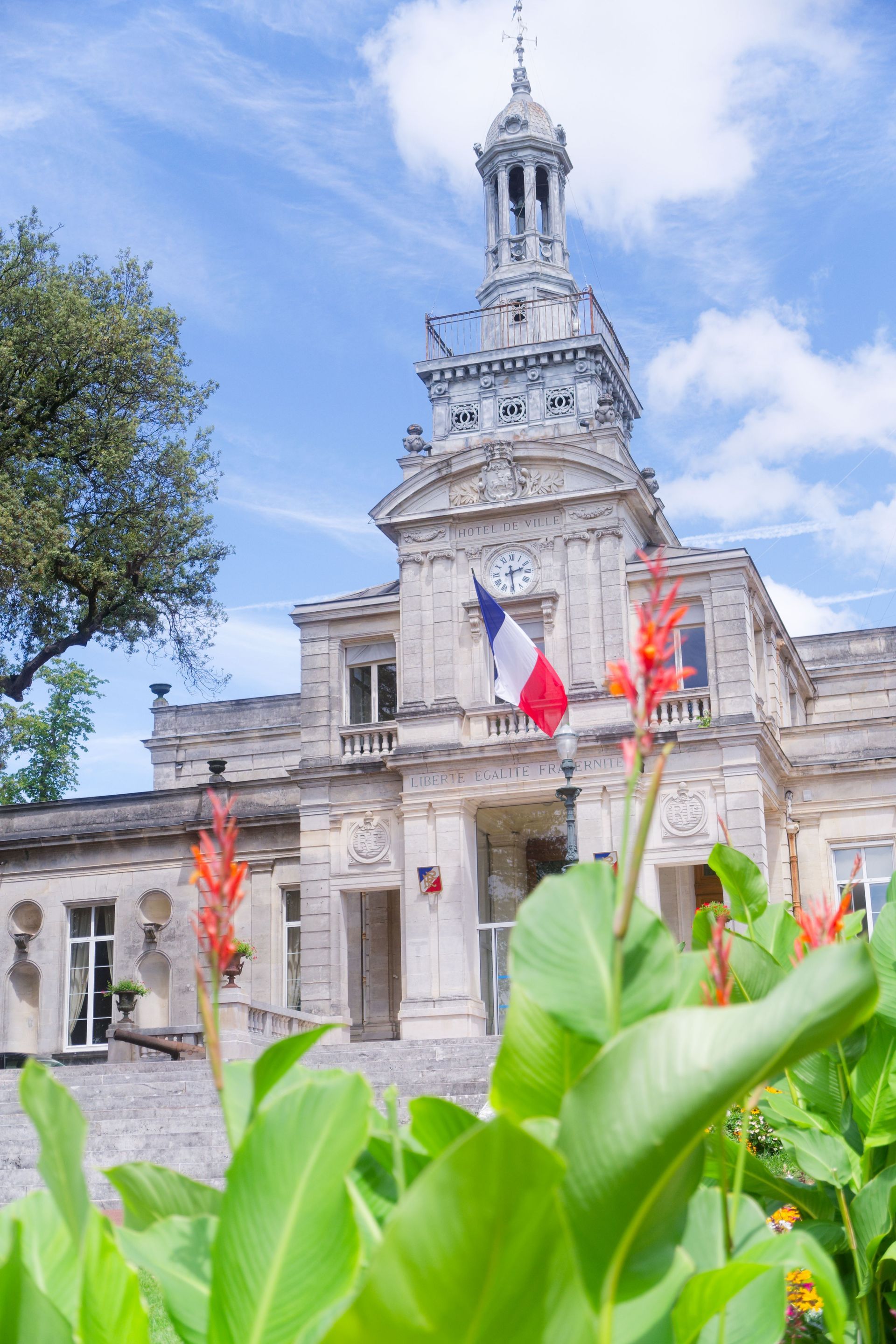 Bâtiment historique en pierre avec une tour ornée derrière des feuilles vertes et des fleurs rouge-orangé sous un ciel bleu