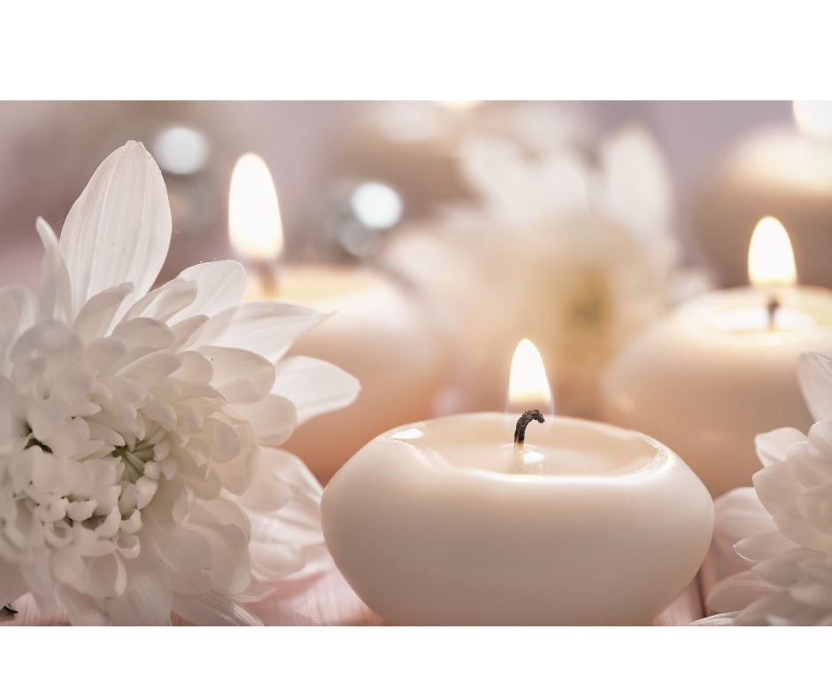 White candles with lit flames, surrounded by white flowers. Soft, blurred background.