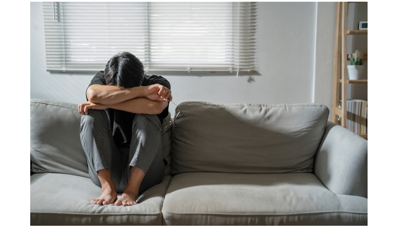 Person sitting on a couch, head in arms, looking dejected. In a brightly lit room near a window.