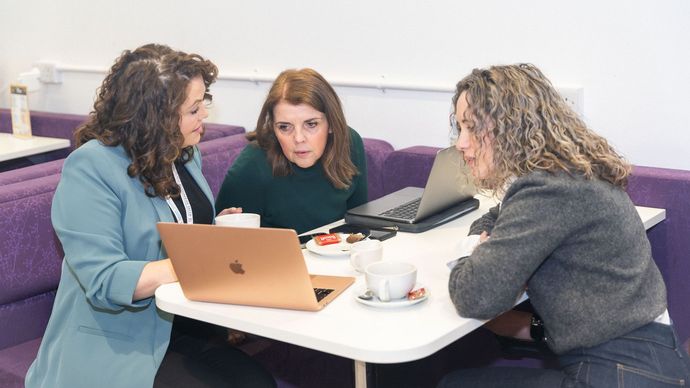 Three women in a cafe, looking at laptops and having a discussion over coffee.