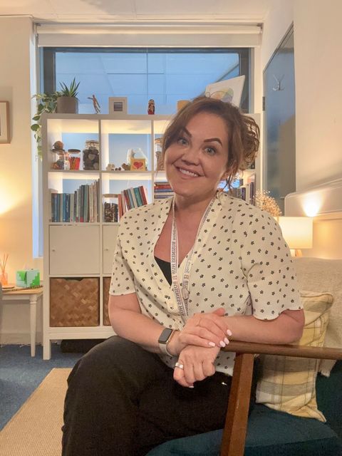 Woman smiling, seated, arms crossed. Wearing polka dot top, sitting in a cozy room with bookshelves and window.