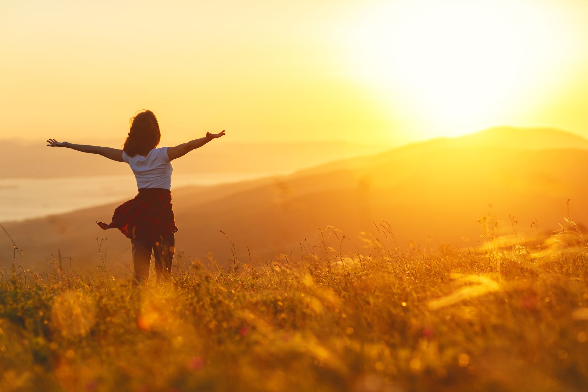 Woman with arms outstretched stands in a field, facing a vibrant sunset.
