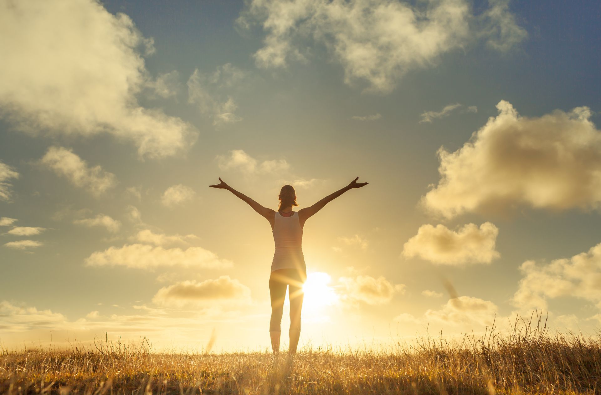 Woman with arms outstretched, standing in a field facing setting sun. Sky with clouds, golden light.