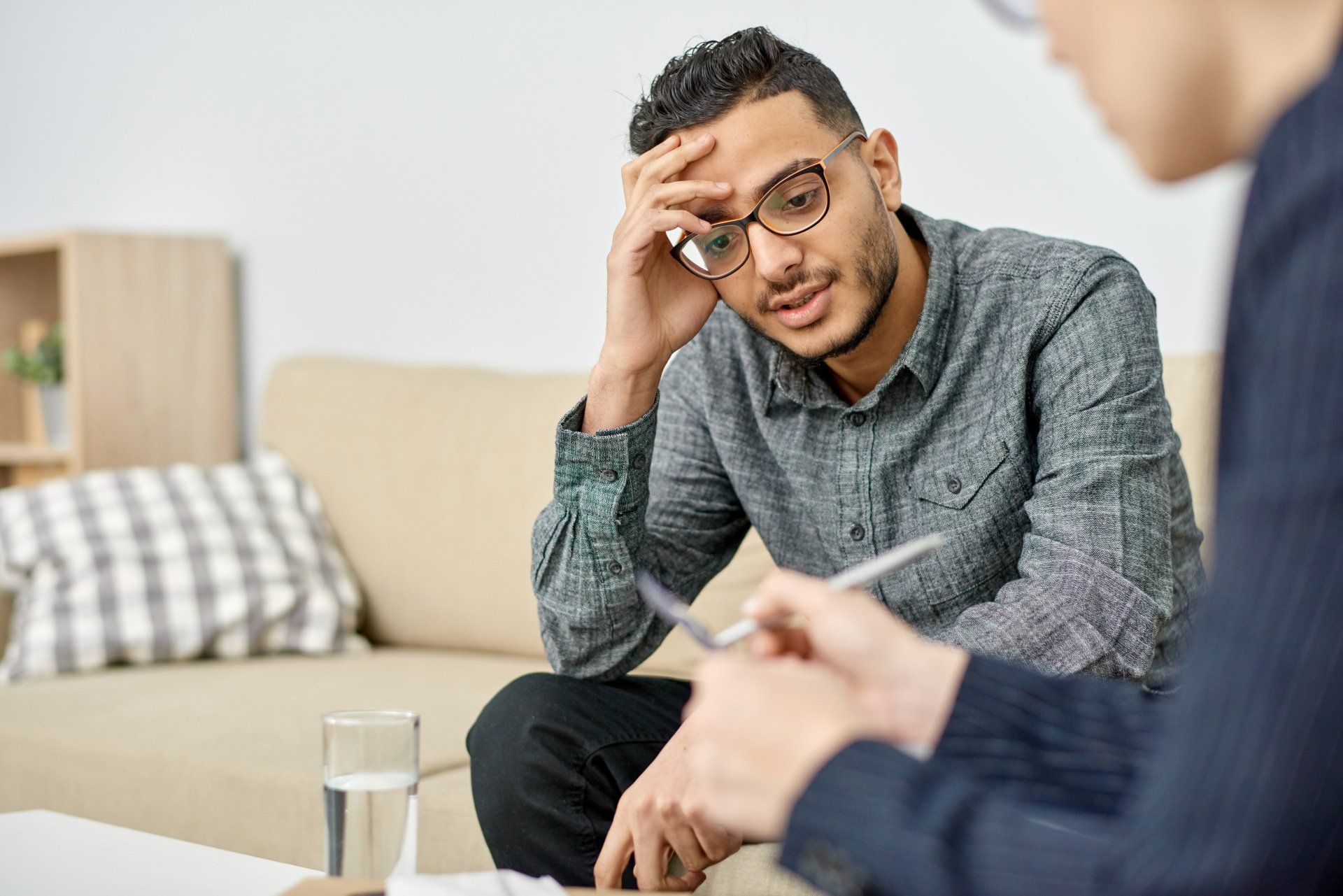 Man in glasses talking, holding his head, with another person sitting opposite, inside a room.