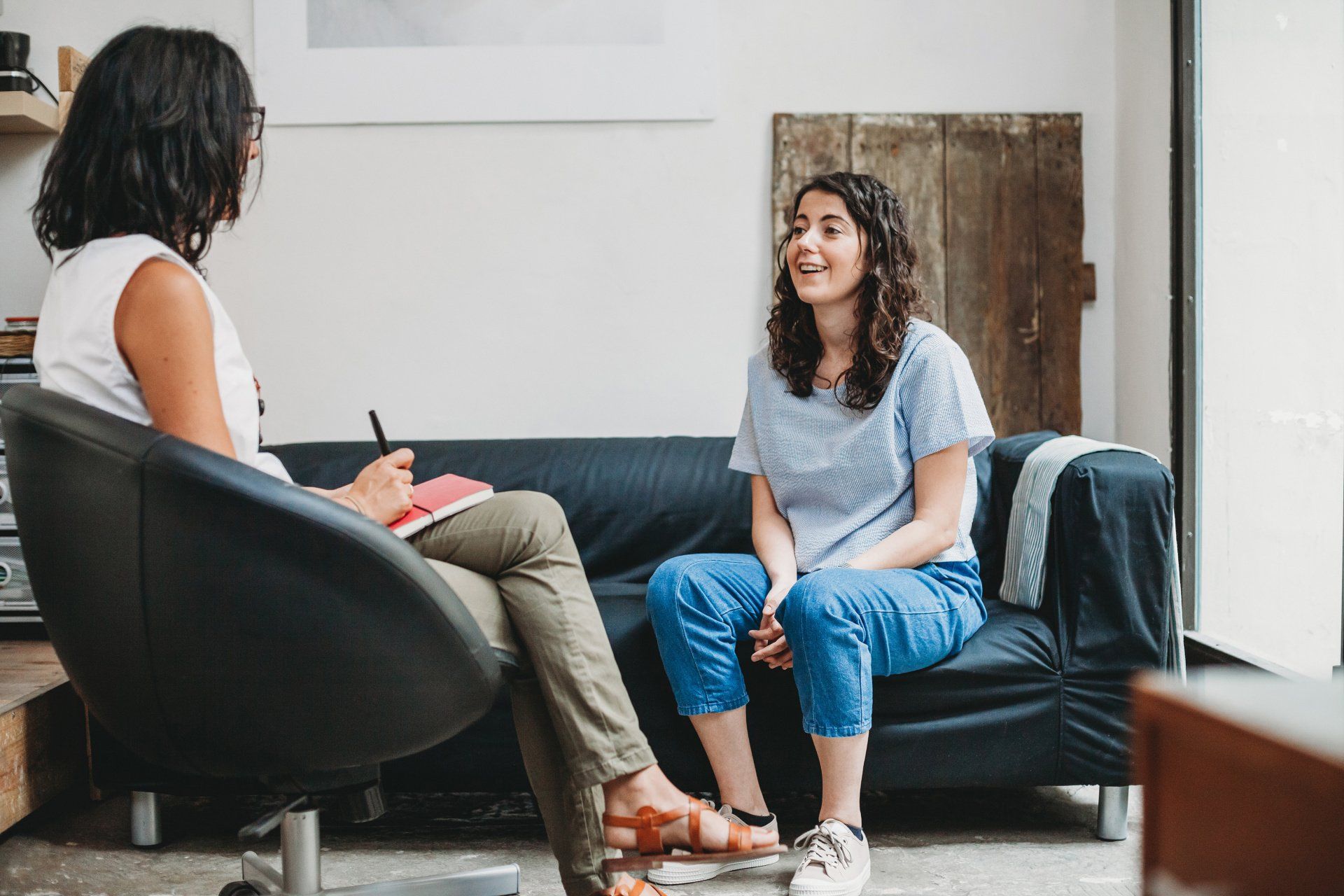Woman on a couch talks to a person holding a notepad in an office setting.