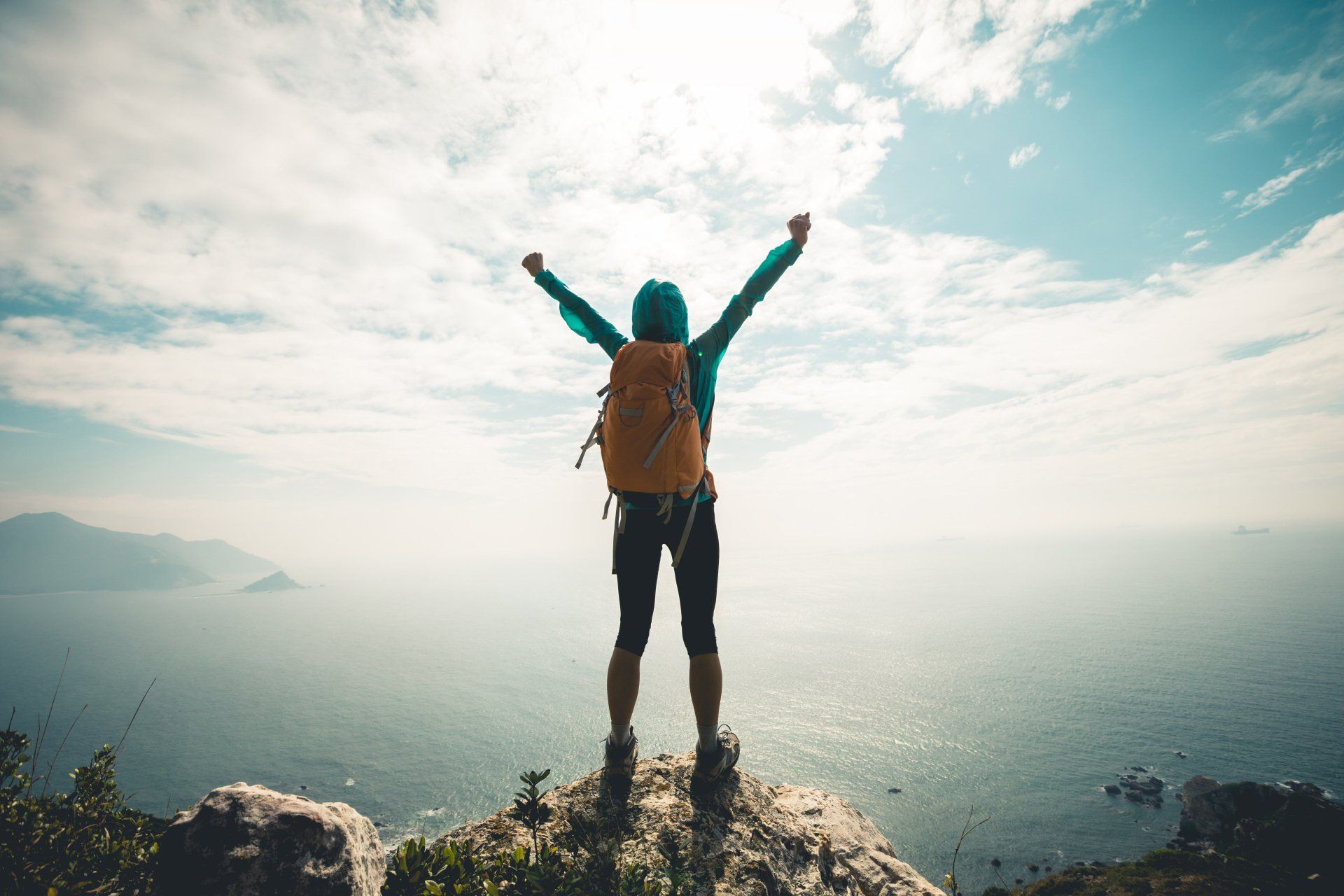 Hiker with arms raised on mountaintop overlooking ocean under cloudy sky.