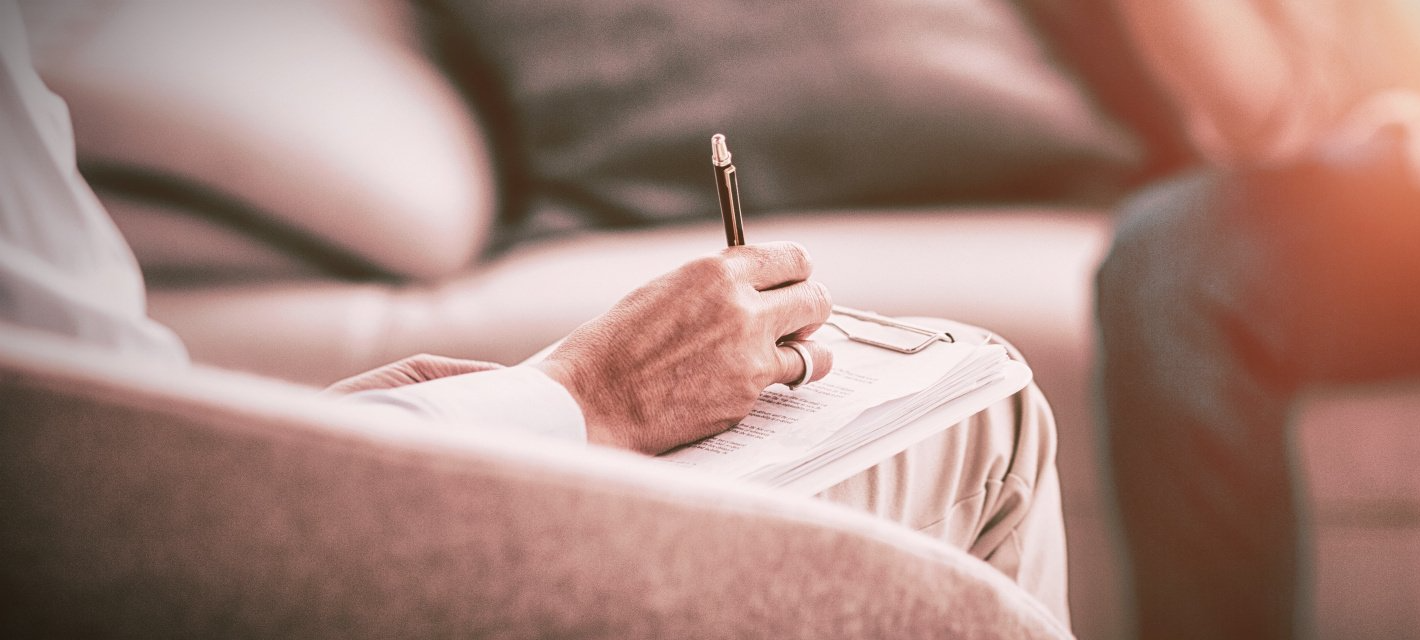 Person writing in a notebook, blurred background of a couch, possibly in a therapy session.
