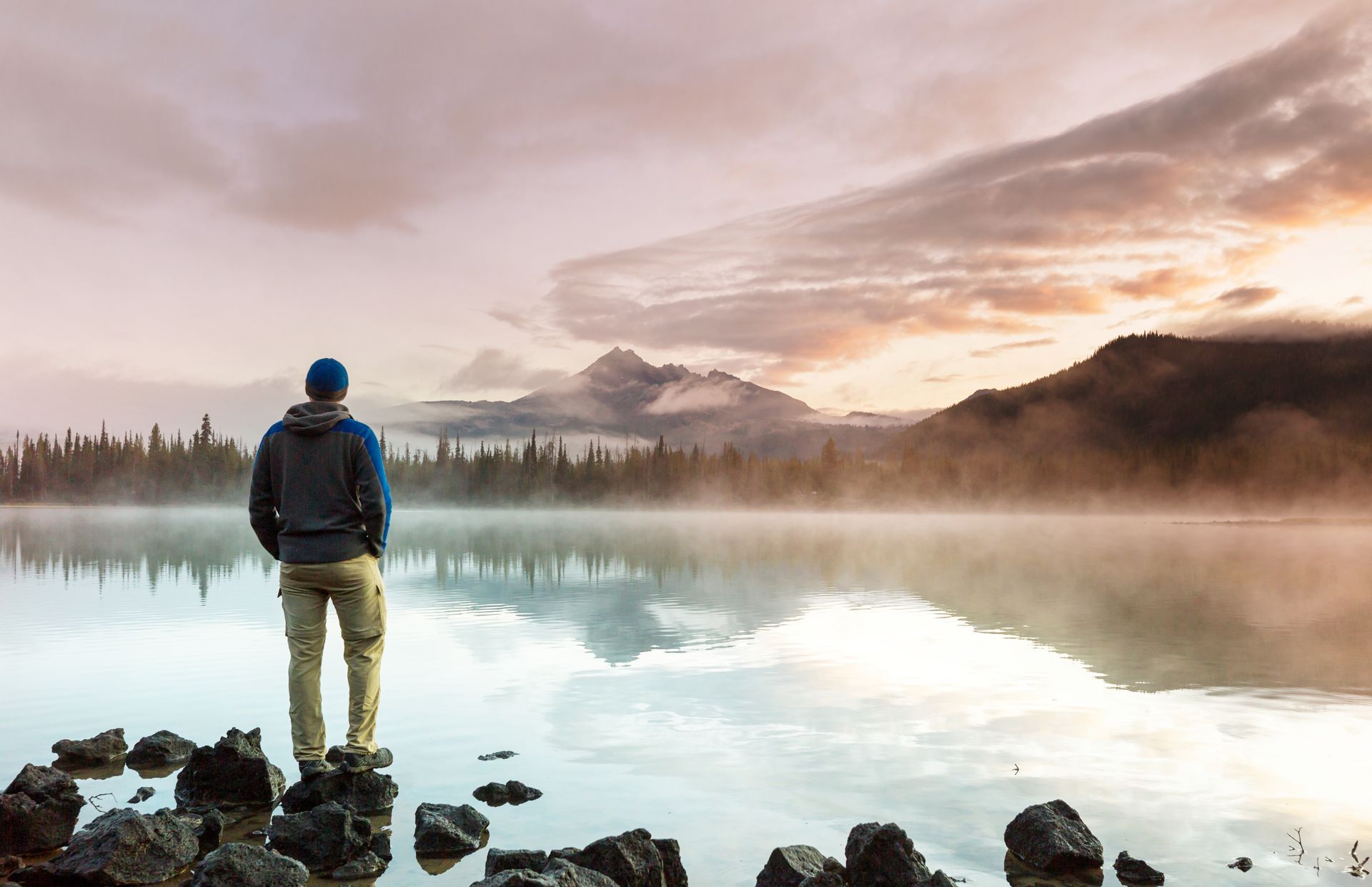 Man standing on rocks, looking at a misty lake reflecting a mountain and sky at dawn.