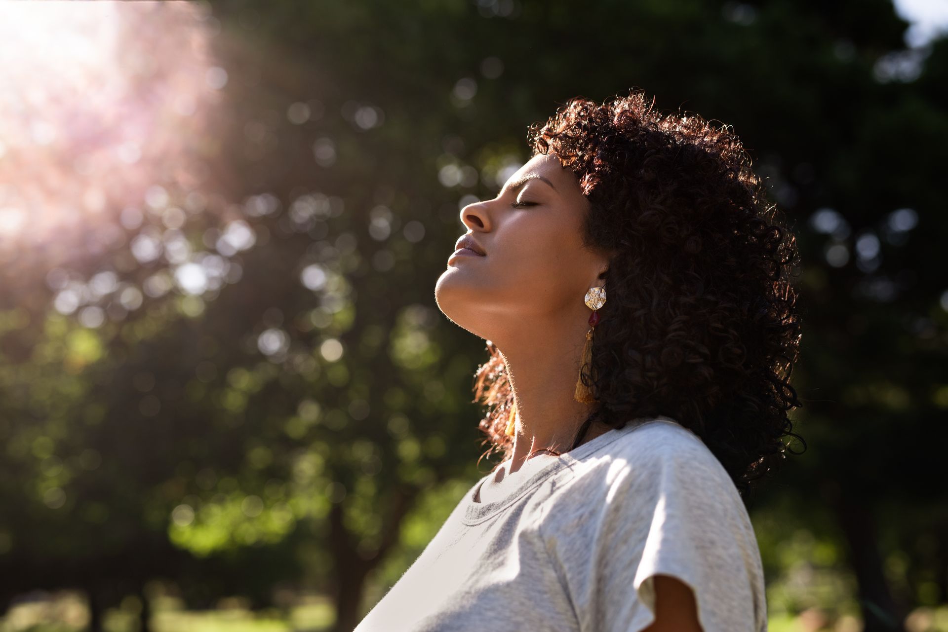 Woman with curly hair, eyes closed, face tilted up, basking in sunlight in a park setting.