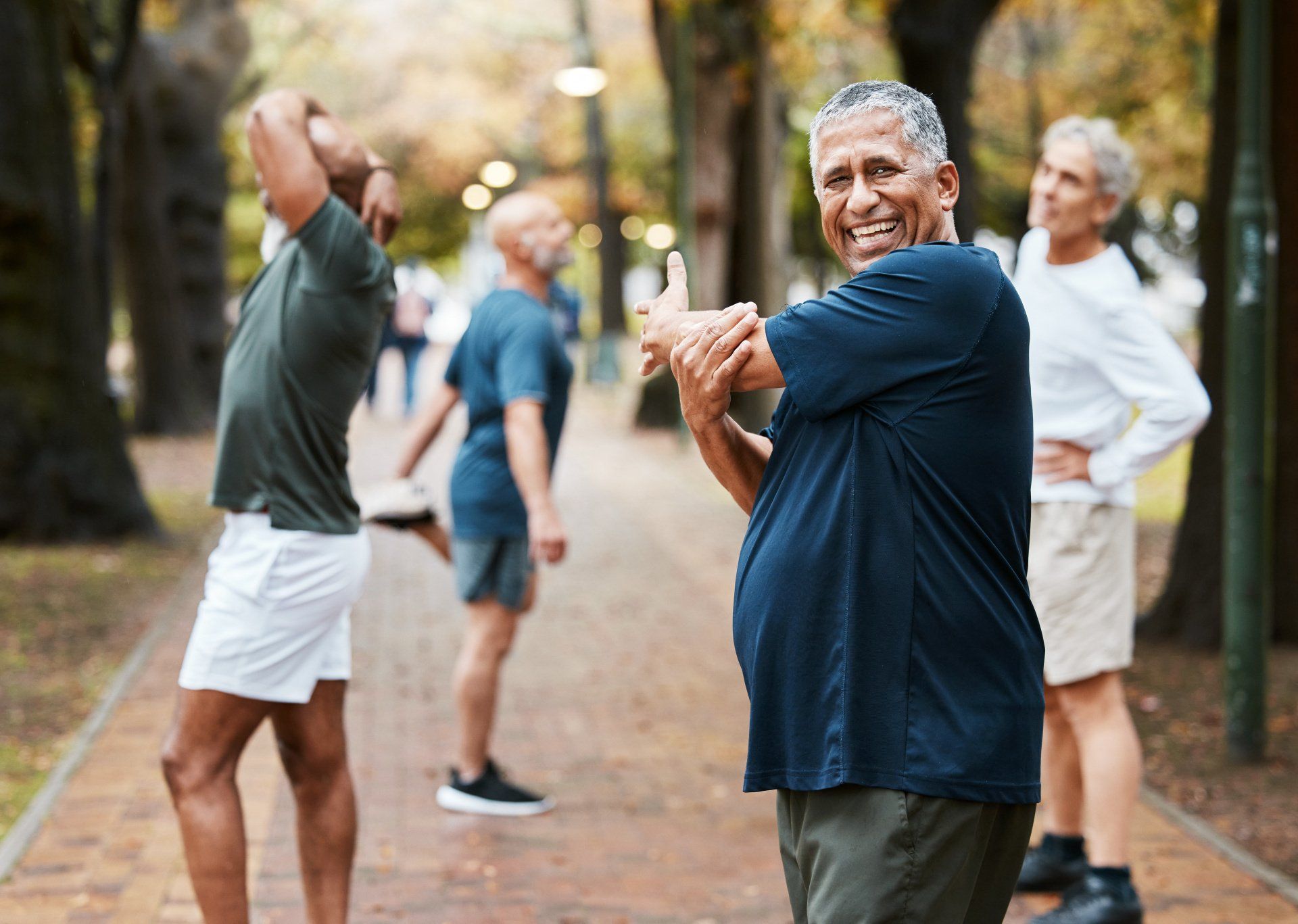 Men stretching outdoors on a tree-lined path. One man smiles at the camera while stretching his arm.