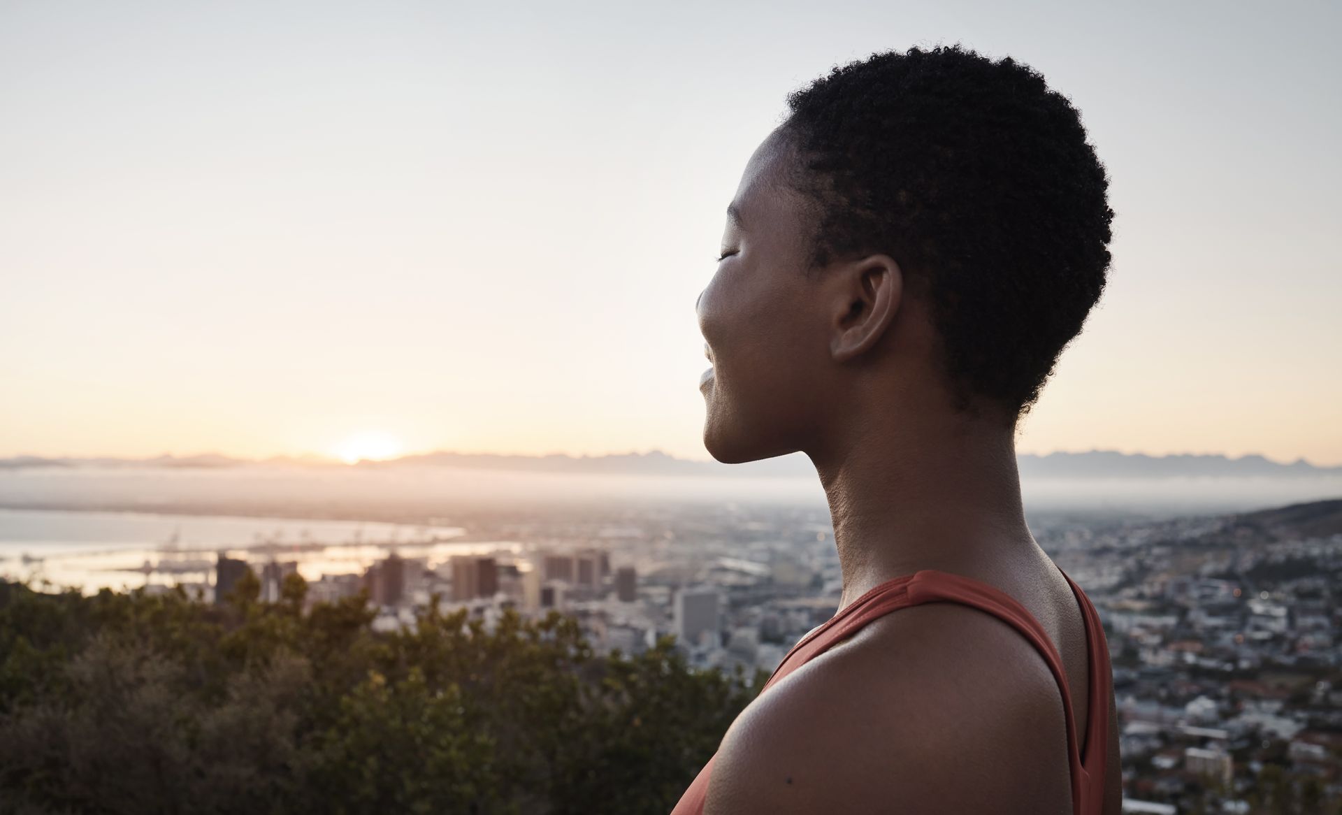 Woman looking at a cityscape from a hillside at sunset; orange top, short dark hair.