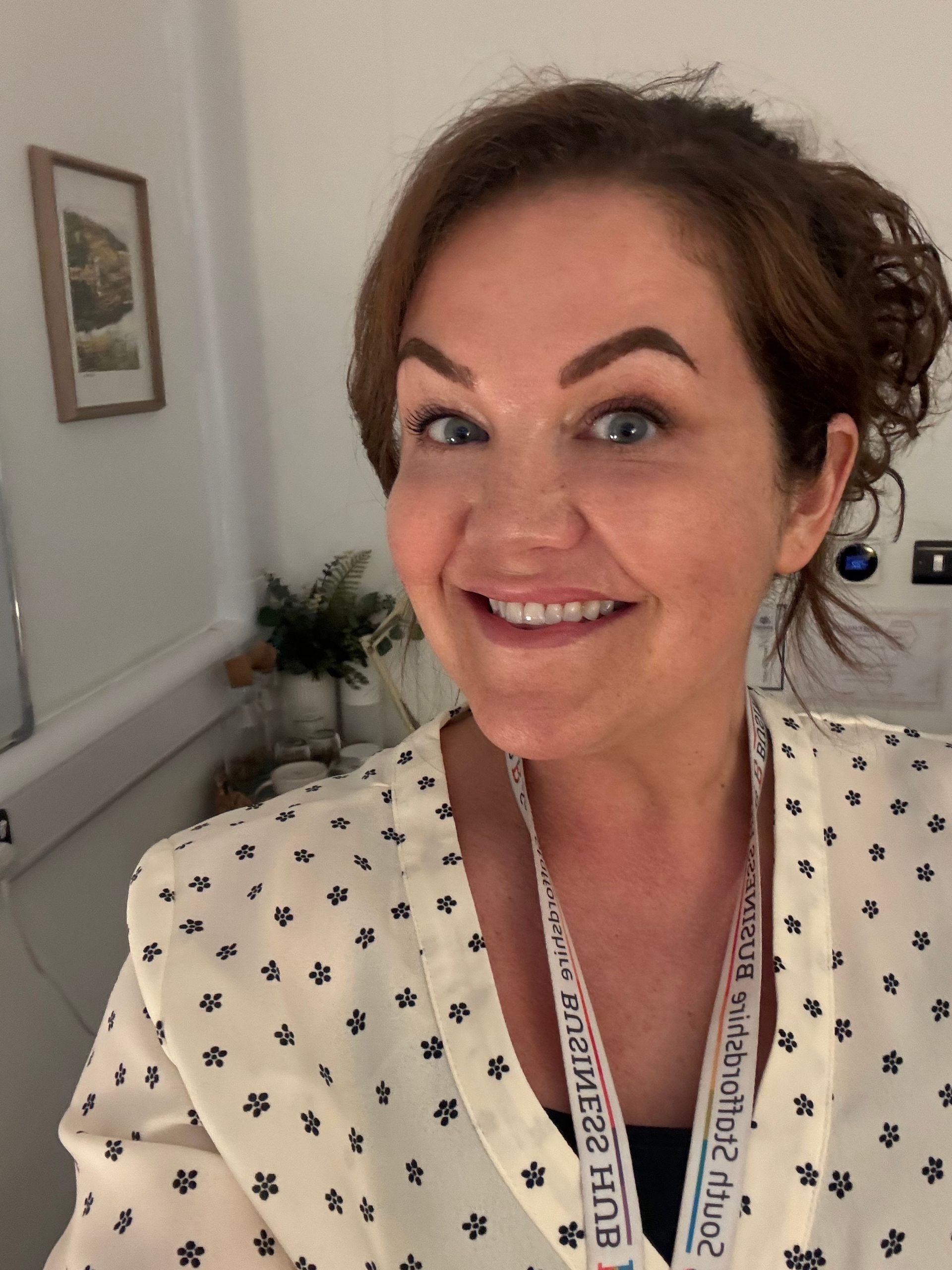 Woman smiles, wearing a patterned shirt and lanyard, in a room with a painting and greenery.