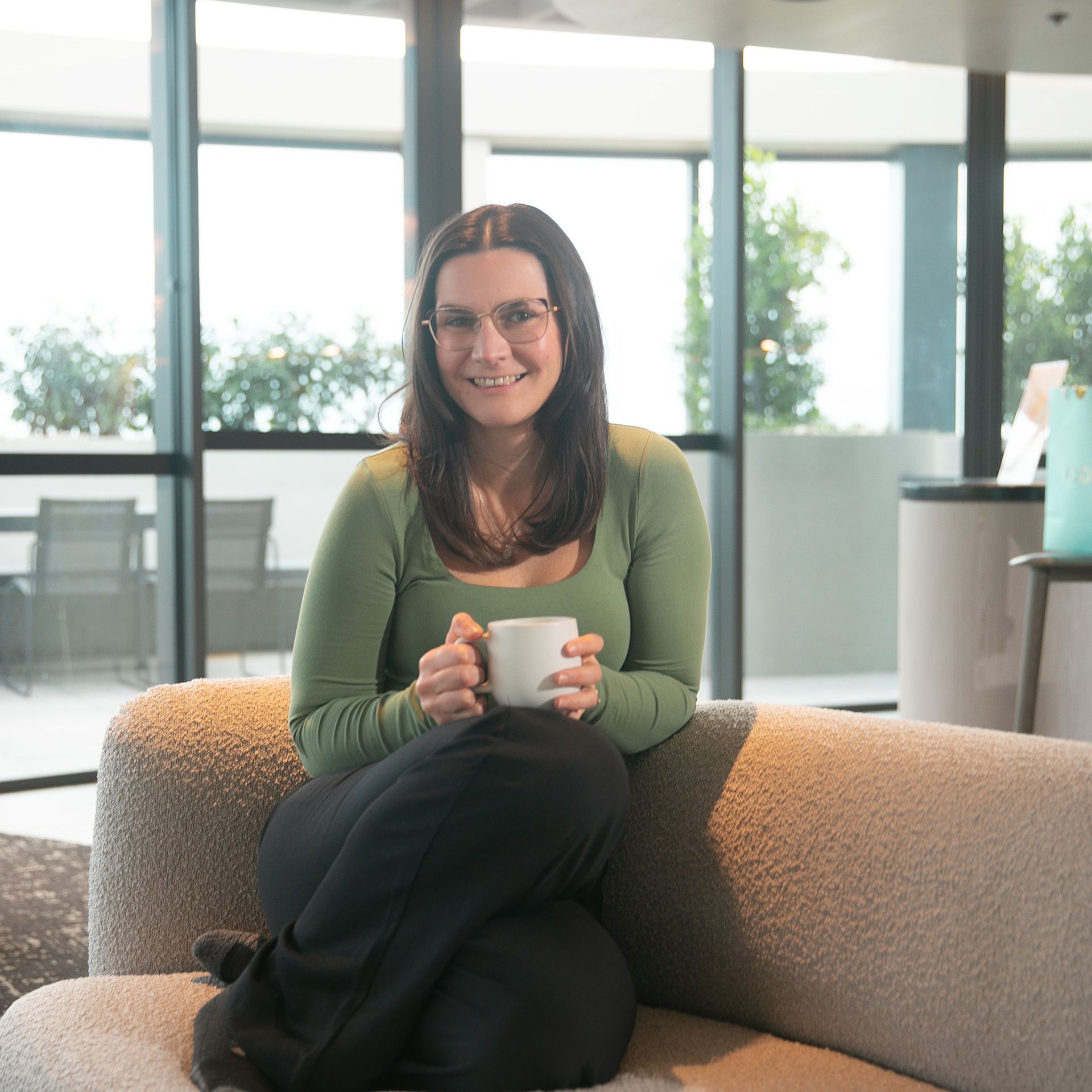 Woman in green shirt, holding a mug, seated on a beige sofa near a window.