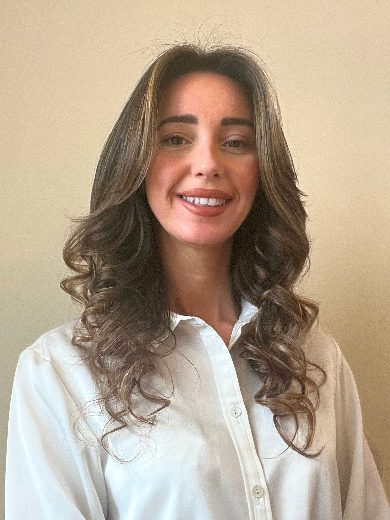 Woman with long curly brown hair wearing a white button-down shirt smiles at the camera.