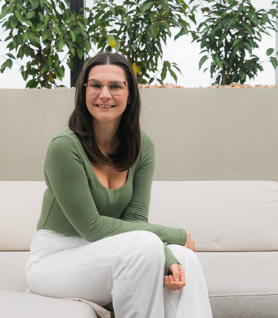 Woman in green shirt and white pants smiles, sitting on a couch. Background includes plants.