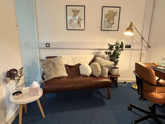 A cozy room with a brown couch, artwork, and a desk. Includes a gold lamp, a small table, and a plant.