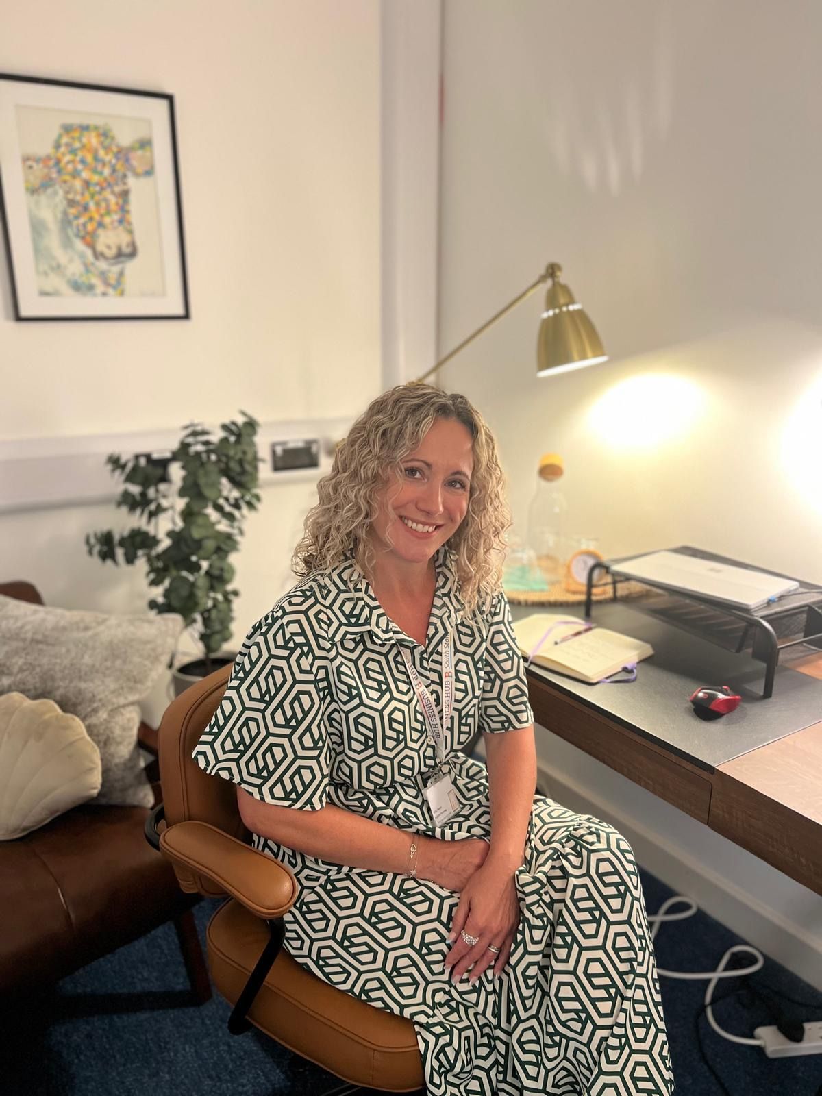 Woman in a patterned dress smiles, sitting at a desk with a lamp, a picture of a cow on the wall.